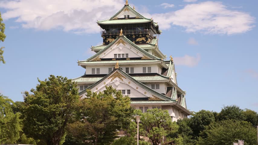 View of the iconic main tower of Osaka Castle in Japan, rising above surrounding trees and showcasing traditional Japanese fortress architecture under a bright sky.