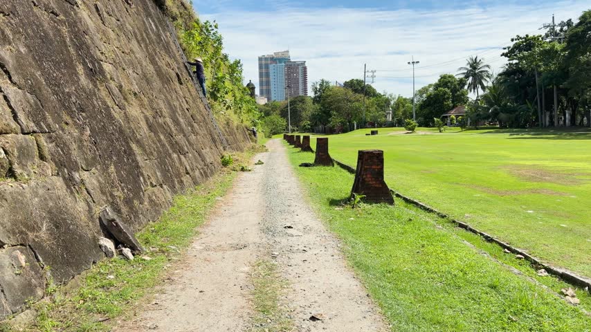 Worker with conical hat climbs long metal ladder leaning against ancient stone wall during routine landscape maintenance at public park with green grass and modern buildings nearby.