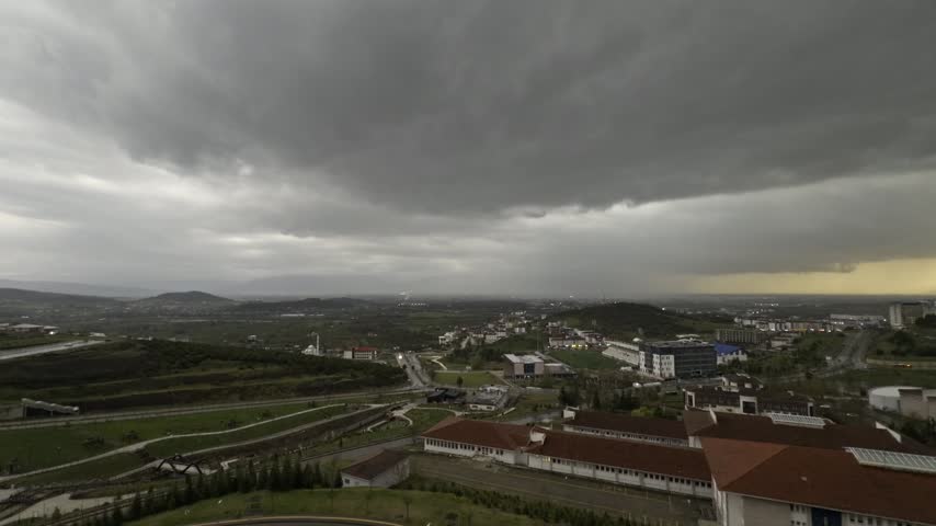 Elevated aerial panorama of long red roofed campus buildings landscaped terraces and a distant town as heavy dark storm clouds roll in and golden sunset light glows on the horizon