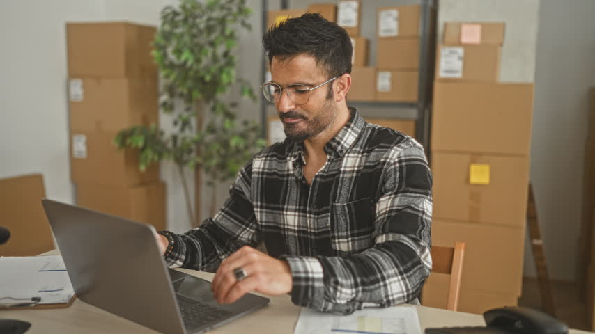 Man with fist raised celebrating over laptop and cardboard parcels in a small business packing area inside a building; entrepreneurial satisfaction.