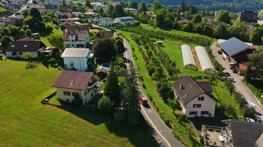 Aerial view over Schwyz, Switzerland, showing hillside homes, orchards, greenhouse tunnels and a farm building with rooftop solar panels beside green Alpine fields.