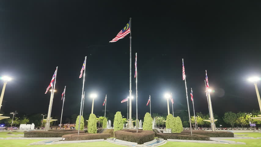 Slow motion low angle shot looking up at Malaysian national flags (Jalur Gemilang) fluttering on towering flagpoles illuminated at night in Dataran Putra, Putrajaya, Malaysia.