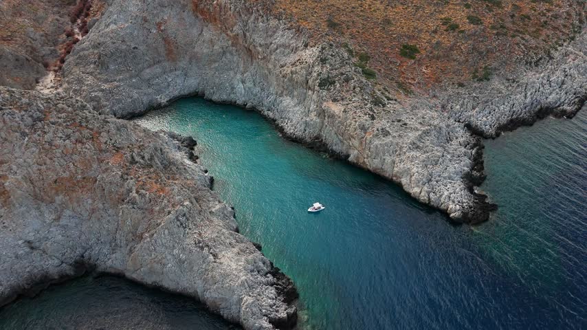 A lone boat floats in turquoise water inside Seitan Limania, Crete, Greece, as a high aerial view follows the narrow inlet between steep rugged cliffs.