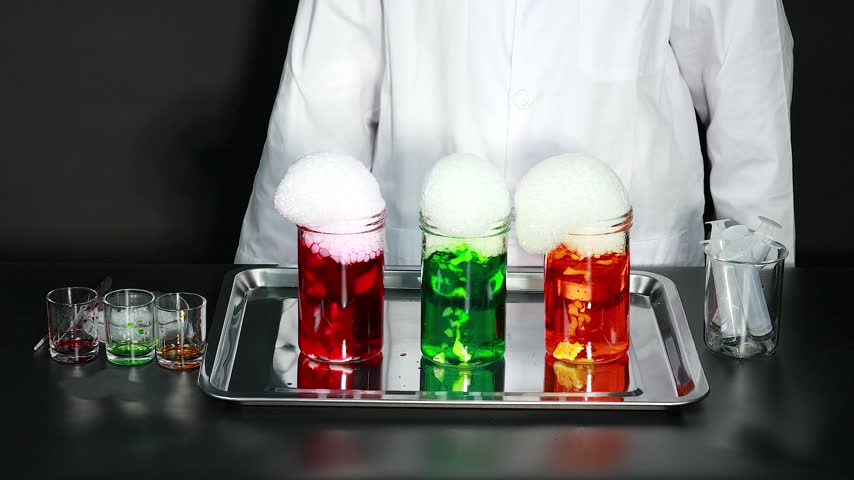 Scientific investigation showcasing chemical reaction with overflowing white foam in three vibrant glass jars placed on metallic tray against black background cinematic studio footage.