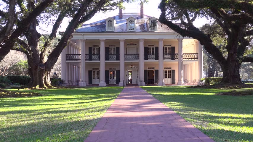 Historic antebellum mansion estate surrounded by massive oak trees with draping Spanish moss, architectural daylight view.