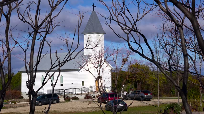 White chapel with steeple set against cloudy blue sky surrounded by autumn trees and parking lot with several vehicles.
