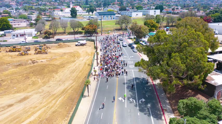 Crowd of protesters marching through suburban neighborhood streets with signs and banners for social justice and equality.