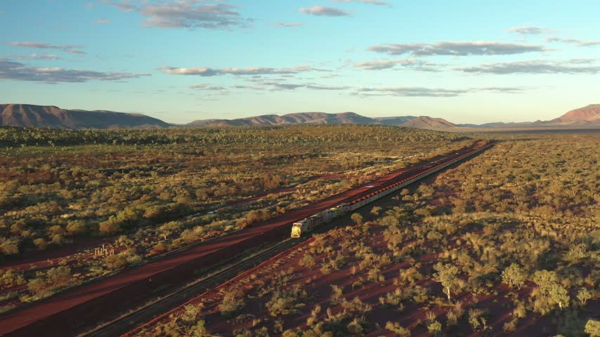 Long cargo train crossing vast Australian desert. Drone tracking shot of red dirt and green shrubbery during sunset.