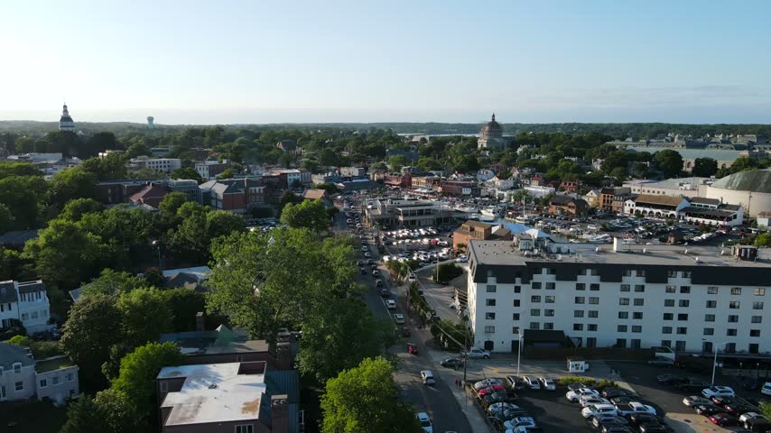 Aerial view of coastal town harbor with docked boats, moving traffic on historic streets, and bright sunshine.
