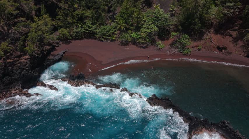 Aerial view of Kaihalulu Beach on Maui Hawaii island with red volcanic sand curving along turquoise water as white surf breaks over dark lava rock reef below lush tropical jungle cliffs.