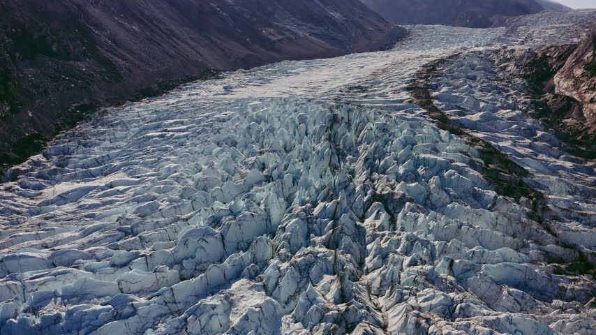 Crevassed Glacier Ice Field Flowing Between Rocky Mountain Slopes In Greenland. - aerial shot
