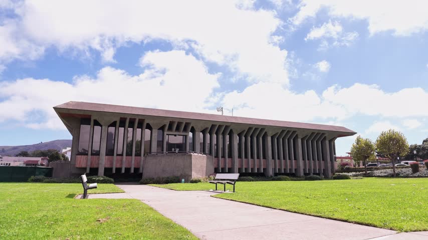 Modernist architecture of the City Hall in Daly City, California, with green lawn and a concrete path leading to the entrance under a blue sky with soft clouds on a sunny summer day