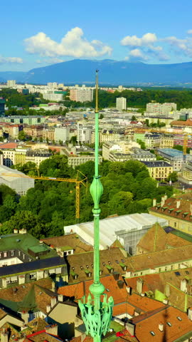 Vertical video. Geneva, Switzerland. St. Pierre Cathedral dominates the Old Town skyline from the highest point of the hill, its distinctive green copper spire and asymmetric twin towers visible above a dense patchwork of historic terracotta rooftops. The cathedral presents a unique architectural blend of Romanesque, Gothic, and Neoclassical styles, the result of centuries of construction, fire, and renovation that began in the 12th century and continued through the Reformation era when John Calvin adopted it as his home church. The aerial perspective reveals the cathedral