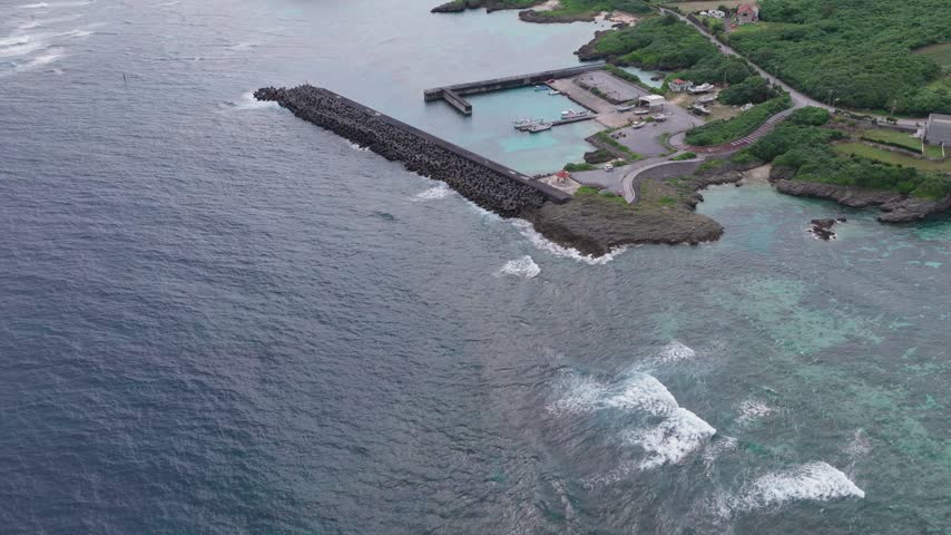 Aerial view of Yuri-Hakuai Fishing Port on Miyakojima Island, Japan, showing harbor structures, waves along the breakwater, turquoise water, and lush green along the shore