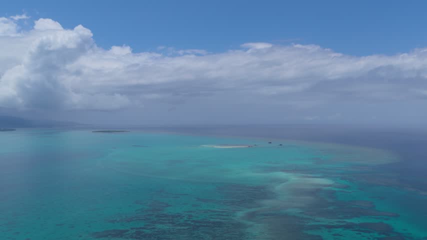 Aerial view of clear blue and turquoise water surrounding a small sandy shore near Phantom Island Hama Island, Okinawa, Japan, beneath a bright blue sky with clouds