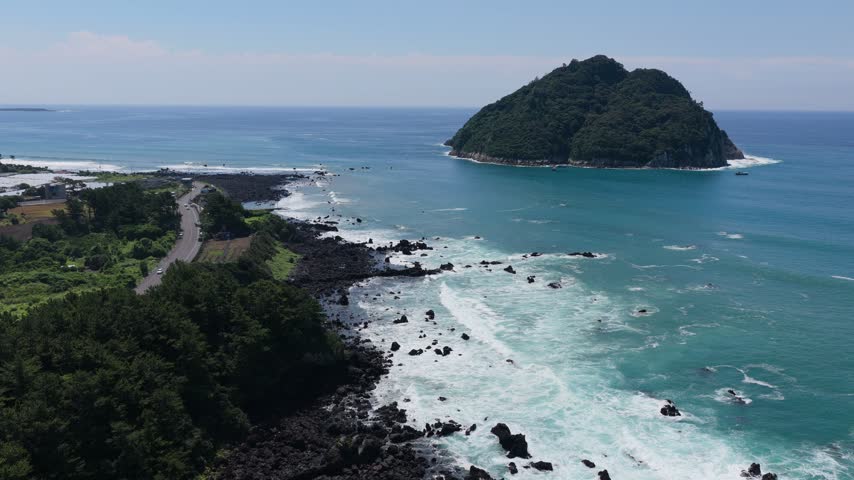 Aerial view of a small forested island near Socheonji, Seogwipo, Jeju Island, South Korea, surrounded by clear blue water, rocky shoreline, and waves under a bright sky