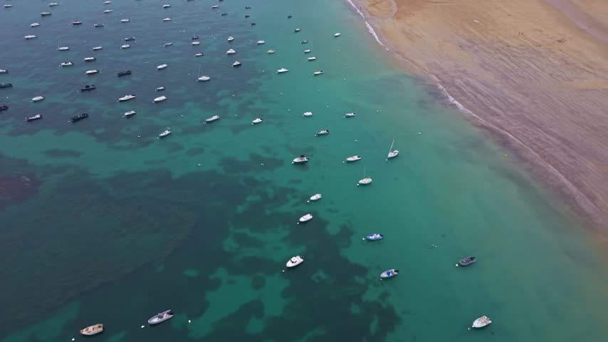 Aerial view of numerous small boats moored in the clear turquoise ocean water near a sandy beach in Saint-Lunaire, France