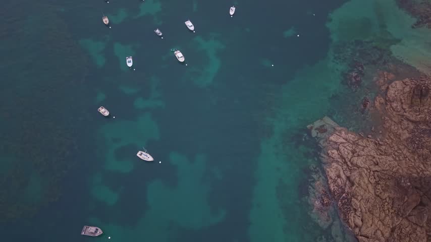 Small boats moored in the clear turquoise ocean water over a rocky seabed in Saint-Lunaire, France. Aerial top-down view
