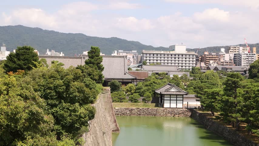 Elevated view of the historic stone walls, moat, and lush gardens of Nijo Castle in Kyoto, Japan, set against the modern urban cityscape