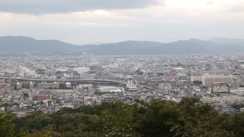 panoramic view over Kyoto from the slopes of Mount Inari near Fushimi Inari Taisha, with the city extending toward distant hills beneath soft atmospheric light