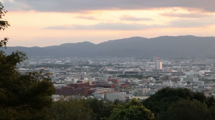 Elevated panoramic view over Kyoto from the slopes of Mount Inari near Fushimi Inari Taisha, with the city stretching toward distant hills beneath soft evening light