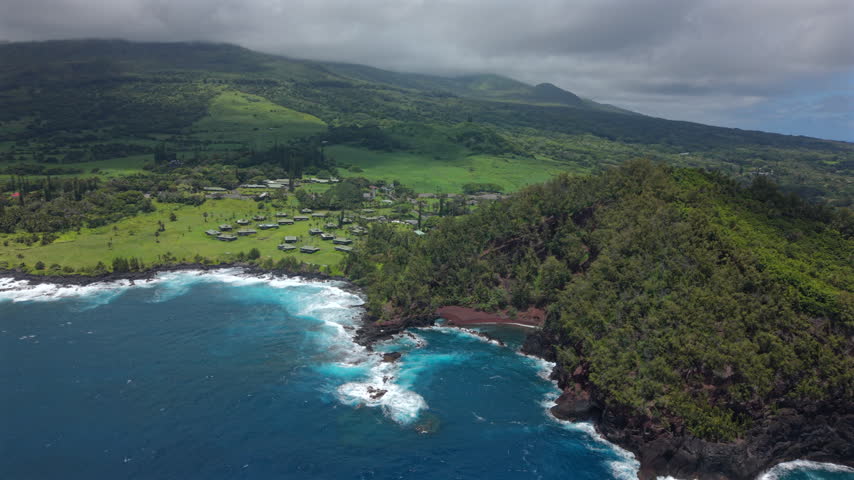Aerial view of Kaihalulu Beach on Maui Hawaii island with red sand cove hidden below green volcanic cliffs turquoise surf crashing on lava rock and misty mountain slopes above the shore.