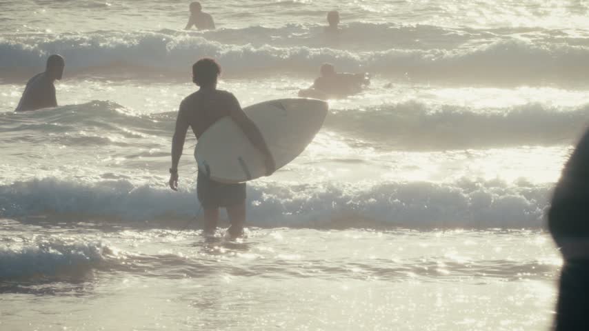Surfers wade through the shallows carrying their boards at Imsouane, Morocco, backlit by sunlight reflecting off the water.