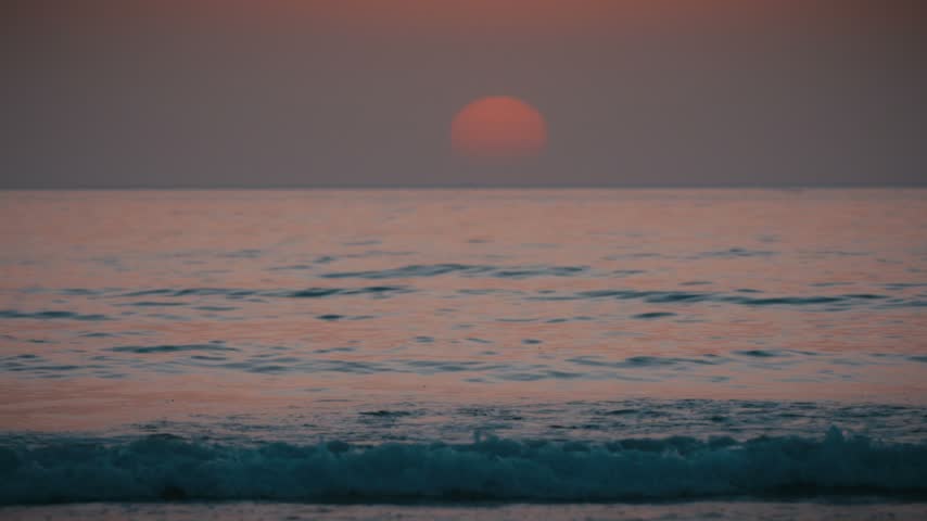 The sun sets over gentle waves on the Atlantic Ocean at Tamraght, Morocco, casting a soft orange glow across the water.