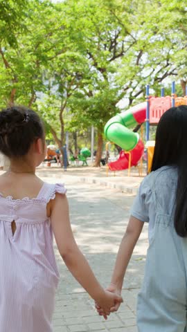 Two young girls hold hands while running toward a colorful playground in Lumpini Park, Thailand.