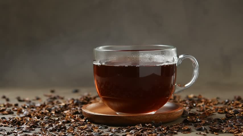 Hand lifts glass cup of cascara tea from wooden saucer surrounded by dried coffee cherry husks.