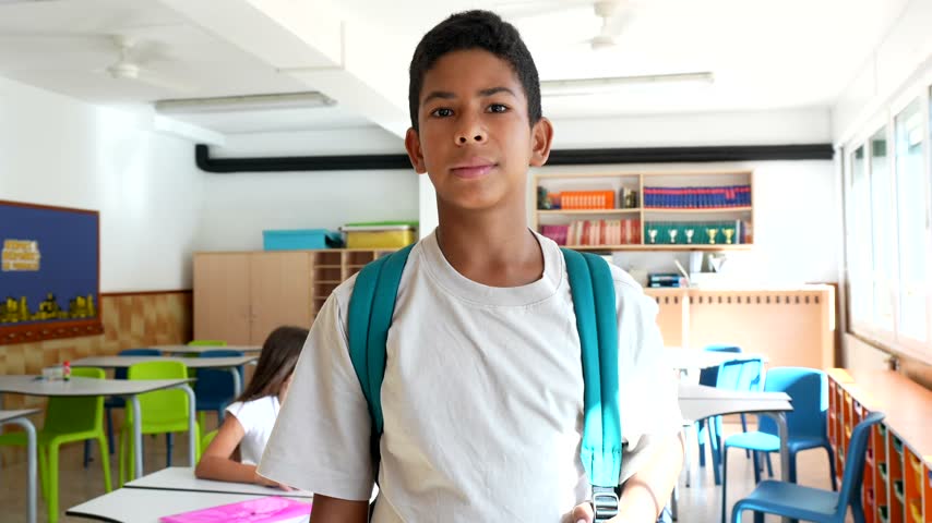 Cheerful elementary school student wearing a backpack standing inside a bright, colorful classroom and smiling confidently at the camera, ready for a new day of learning and education