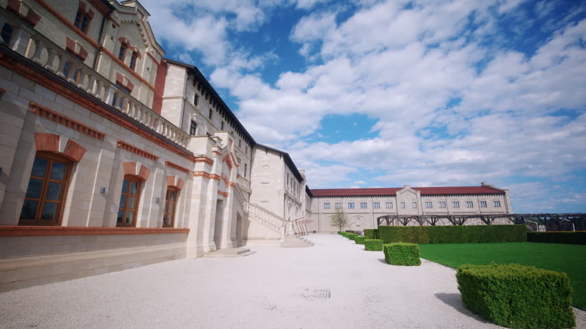 Wide view of the elegant courtyard at castle winery in Moldova, featuring historic stone architecture, arched windows, landscaped hedges, and a bright blue sky with clouds