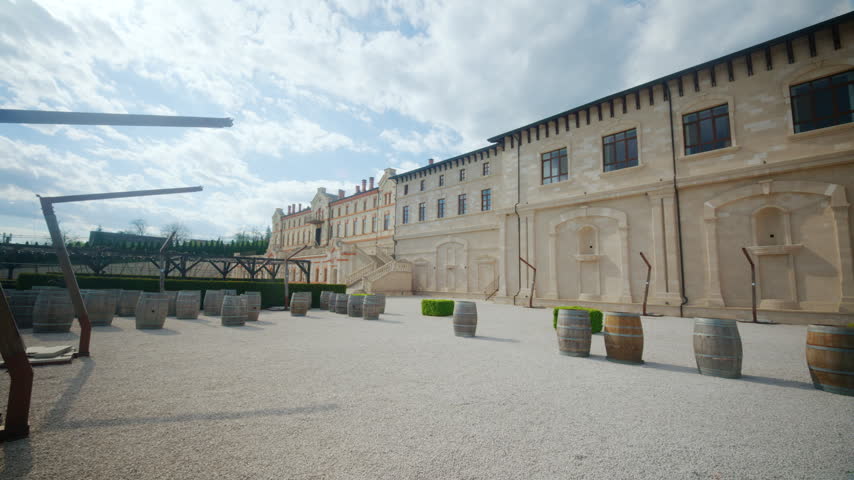 Wide courtyard view at winery castle in Moldova, showing historic winery buildings, stone facades, wine barrels, gravel pathways, and architectural staircases