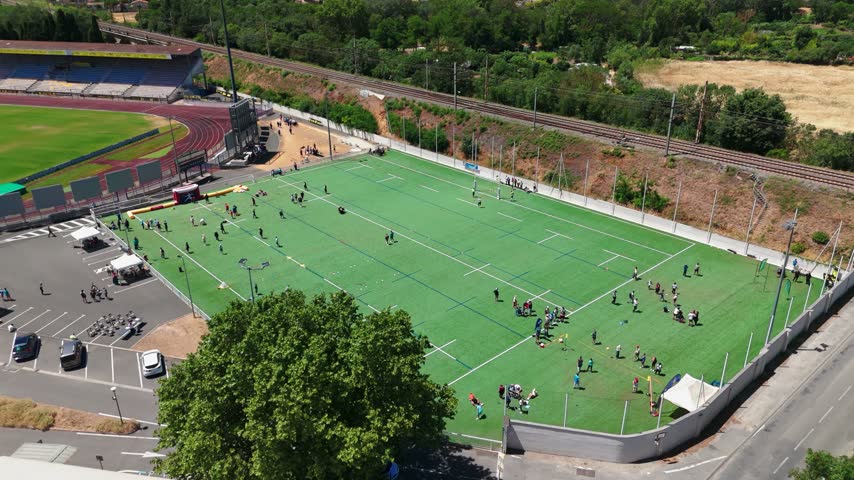 People playing on a green sports field and rugby pitch next to a stadium and railway line in southern France.