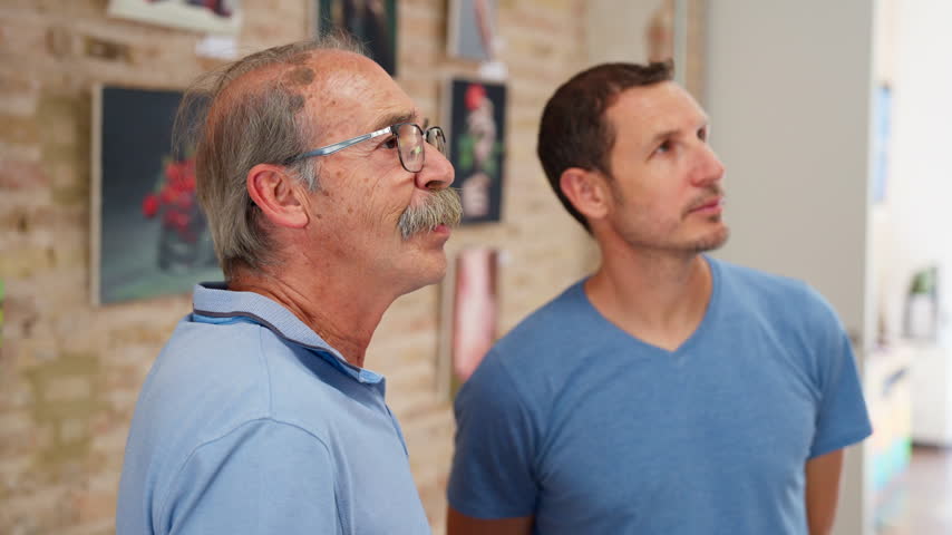 A senior man and a young man stand side by side looking up at paintings displayed on a gallery wall, Slow Motion.
