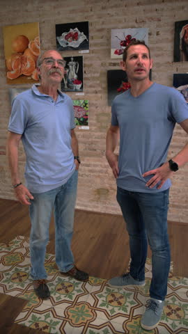 A senior man and a young man stand side by side looking up at still life fruit paintings in an art gallery, Slow Motion.