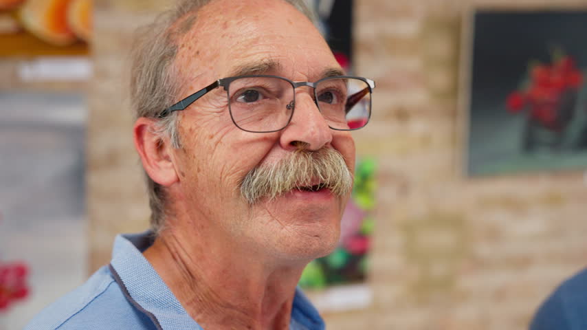 A close up of a senior man with glasses and a mustache talking animatedly inside an art gallery, Slow Motion.