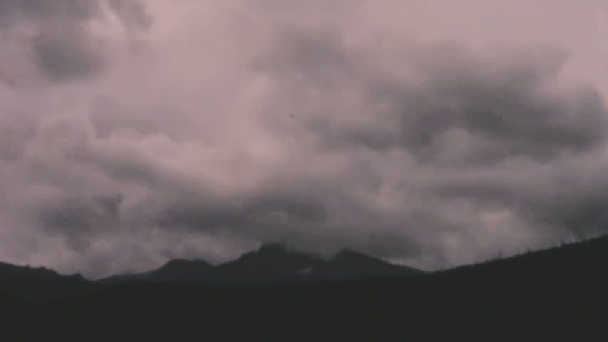 Dark silhouetted ridge leads to a distant Rocky Mountain peak in Colorado. The central mountain is partially obscured by thick grey clouds under an overcast sky archival footage.