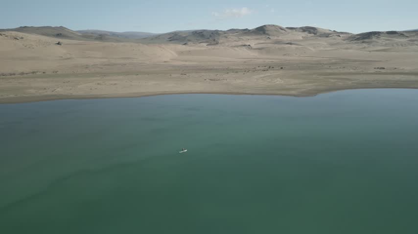 Aerial orbit shot of a woman on a sup board inside an emerald desert oasis lake in Mongolia under a clear blue sky, flat log profile