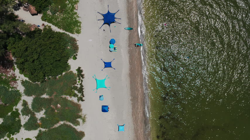 Blue and teal shade tents line a sandy beach while a few people relax near the shore and swim in gentle waves.