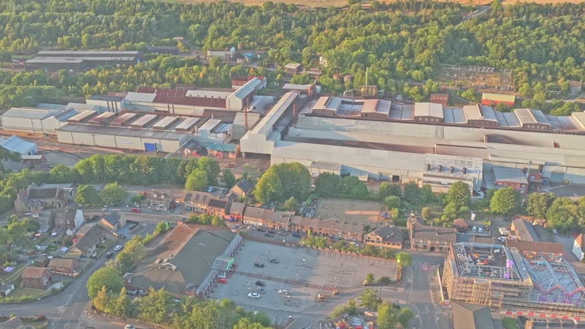 Stocksbridge steelworks extends beside residential streets and wooded hills in Upper Don valley, South Yorkshire, England, with large production halls, drone forward flyover.