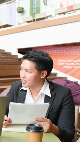 Young Asian professional in suit reviews papers and speaks during casual business discussion.