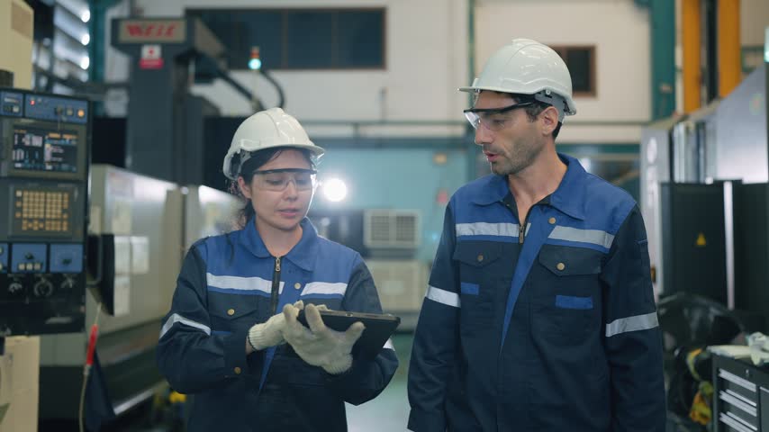 Two workers inspect machinery in a factory. They wear hard hats and safety glasses. One holds a tablet, the other gestures toward equipment