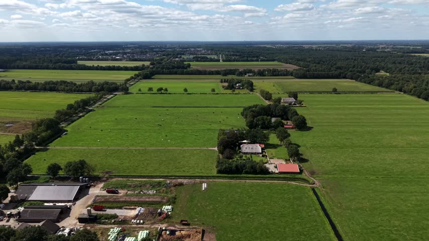 Patchwork green pastures spread around white farmhouse with red barn in rural Pennsylvania, grazing cattle dot open fields beneath scattered summer afternoon rain clouds. Aerial drone wide shot