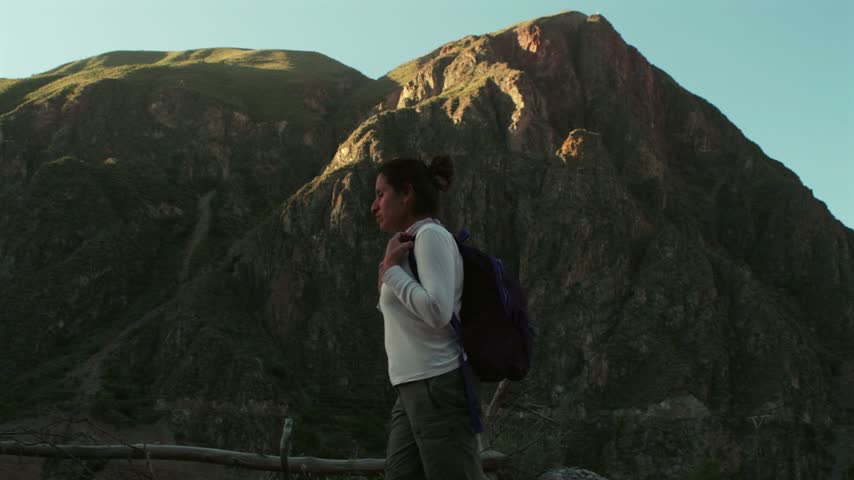 A female traveler walks and gazes at the immense mountains of the Peruvian Andes against a beautiful sunset.