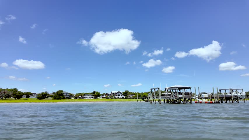 A sweeping pan captures the Hampstead waterfront from a fast-moving boat, with coastal homes, docks, marshy shoreline, and blue summer skies passing across the horizon.