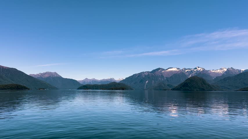 Low boat level view across the calm blue water of Lake Manapouri to bush covered islands and the snow streaked Fiordland mountains beyond, New Zealand. Wide shot