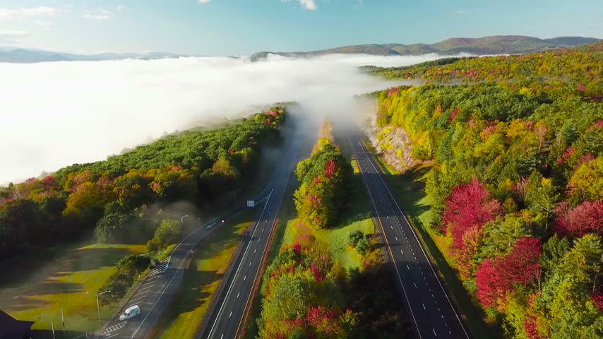 Aerial video showing cars traveling on highway surrounded by colorful autumn trees, morning fog hangs low over distant mountains.