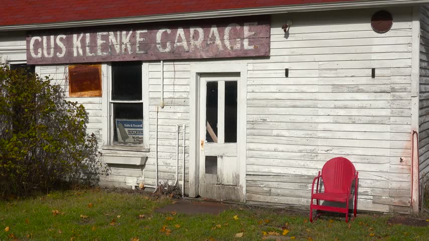 Static shot of historic white wooden garage building with weathered sign and bright red lawn chair on autumn grass.