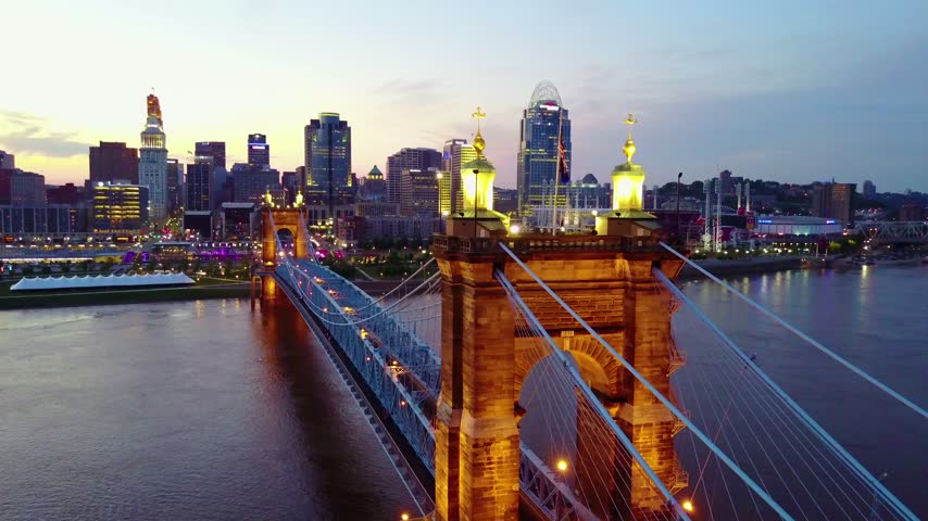 Drone flies over suspension bridge at dusk, city skyline backdrop, lights on bridge and buildings.